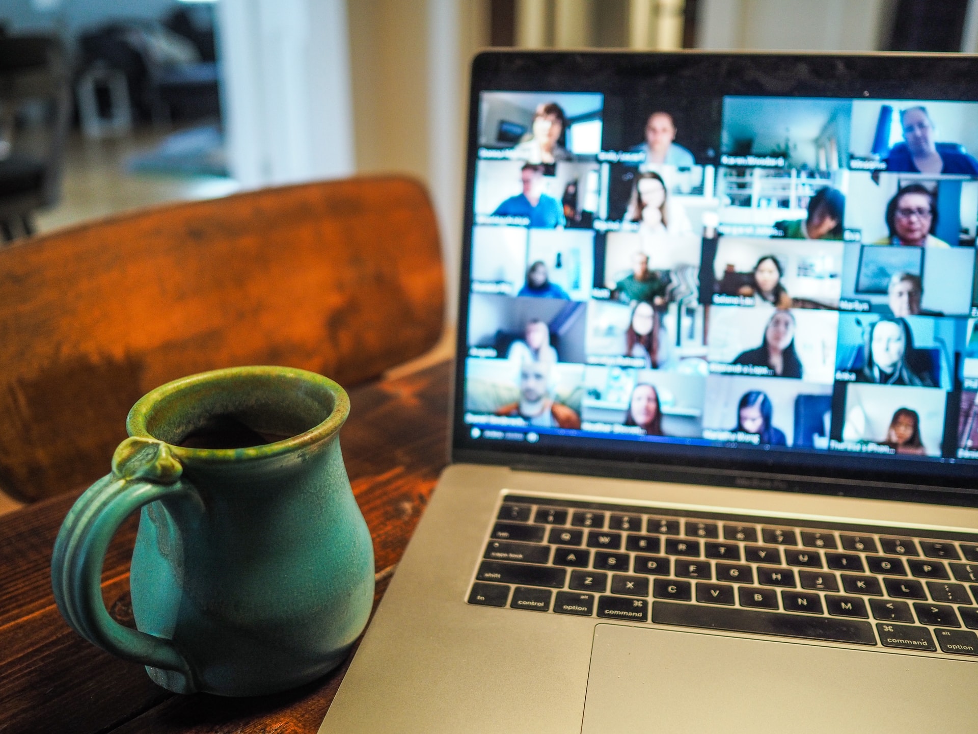 On a dark wooden table is blue mug & a laptop at an angle displaying the many faces of people attending a video teleconference meeting. | Photo: Chris Montgomery via Unsplash 